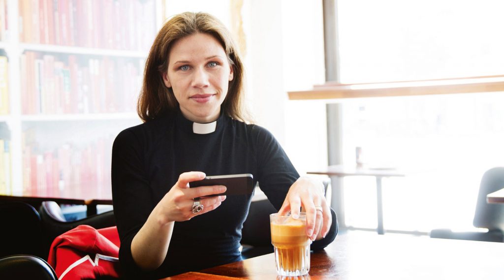 Kvinnlig präst med kaffekopp och mobil tittar in i kameran. Foto Alex & Martin/IKON.